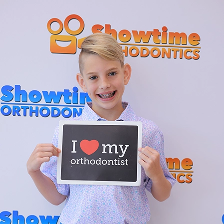 A young boy smiling while holding a sign that says, I heart my orthodontist.